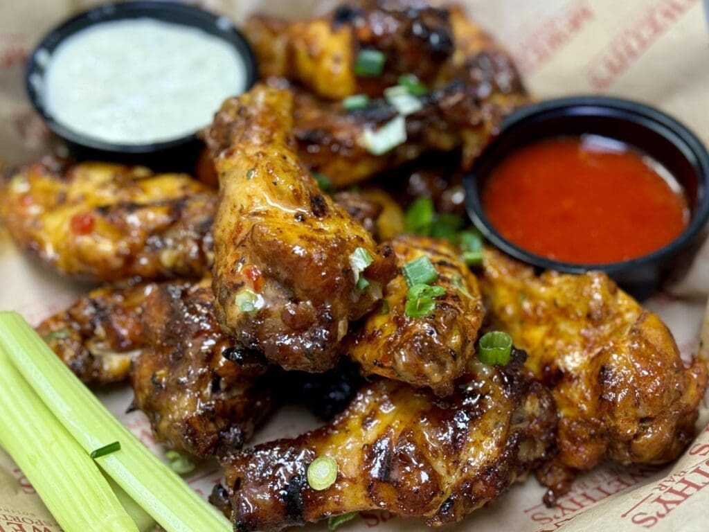 A close-up of Chef Bobby's SWEET CHILI SRIRACHA wings with dipping sauces and celery at McWethy’s Sports Bar.