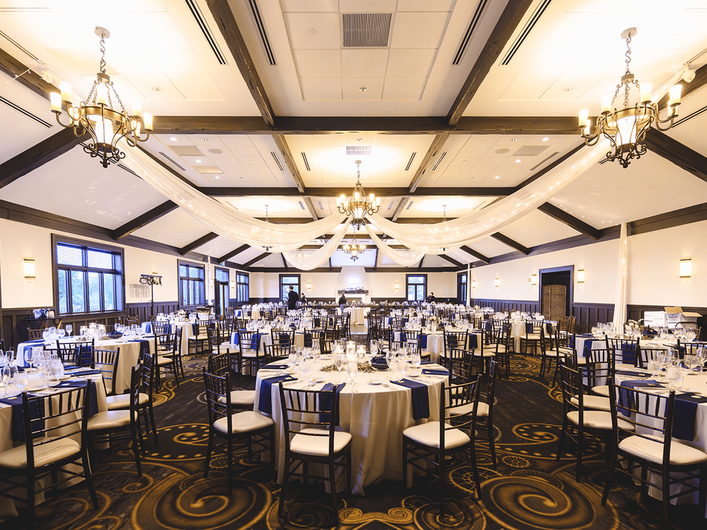 Elegant banquet hall at Mistwood Golf Club set for a wedding reception with white linens, navy accents, and chandeliers beneath vaulted wood-beam ceilings.