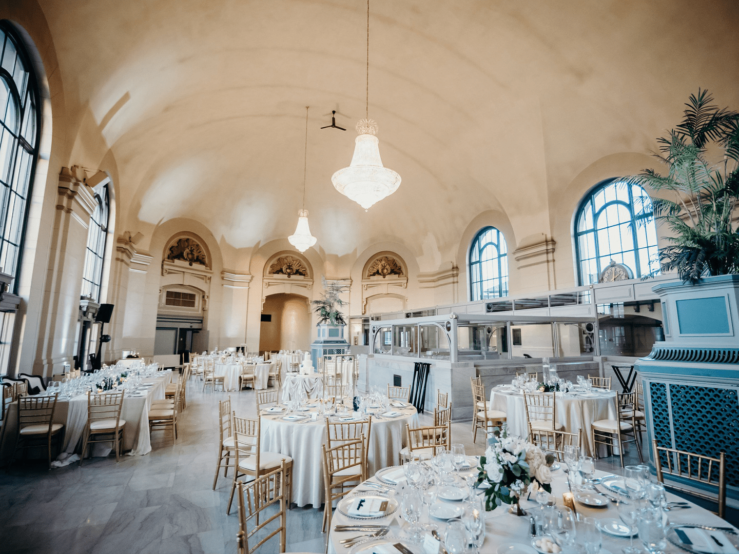 Elegant wedding reception hall inside The Grand Ballroom at Joliet Union Station with gold chairs, round tables, white linens, and crystal chandeliers under a vaulted ceiling.